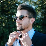 Fashionable man adjusting his suit and sunglasses outdoors on a sunny day.